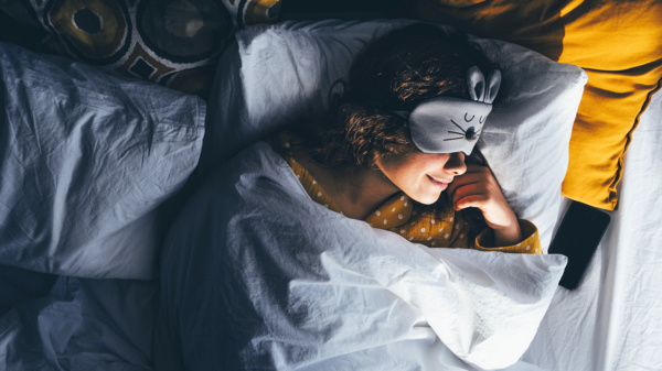 Kobieta w kręconych włosach leży w łóżku z opaską na oczach uśmiechnięta i zadowolona z przyjemnego snu