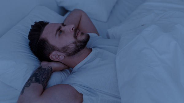 Młody mężczyzna z gęstym zarostem leży w białej pościeli i rozmyśla