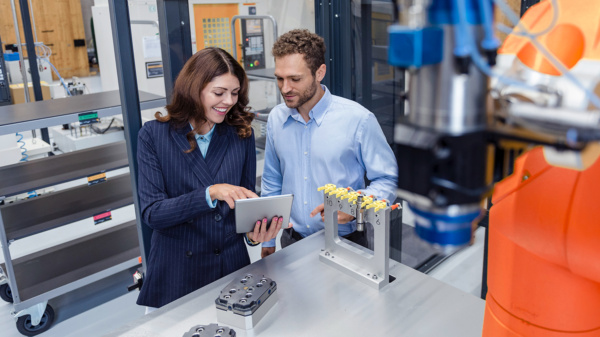 kobieta i mężczyzna znajdują się w pracowni robotycznej. Kobieta ubrana w ciemną marynarkę przedstawia coś mężczyźnie w jasnej koszuli na tablecie.