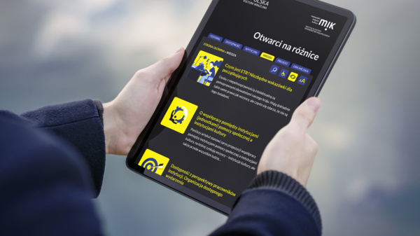 Tablet trzymany w dłoniach, a na jego ekranie wyświetla się strona poświęcona dostosowaniom dla osób z niepełnosprawnościami.