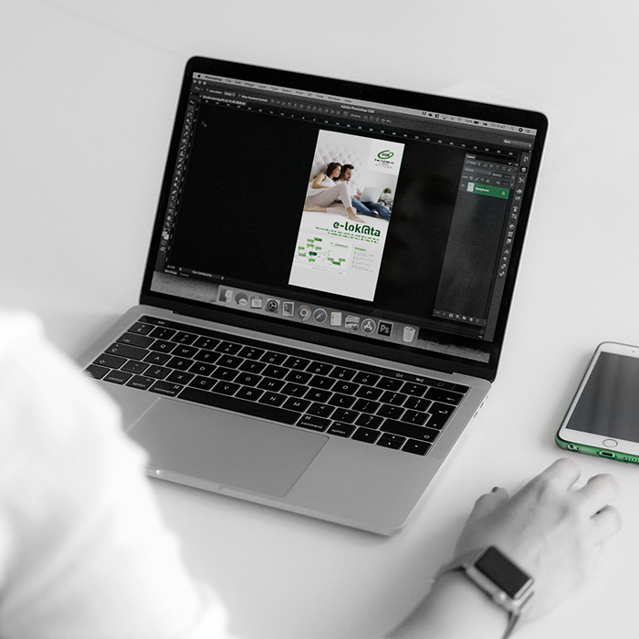 Osoba pracująca na laptopie, na ekranie widoczny projekt ulotki reklamowej „e-lok@ta” Banku Spółdzielczego, obok leży smartfon