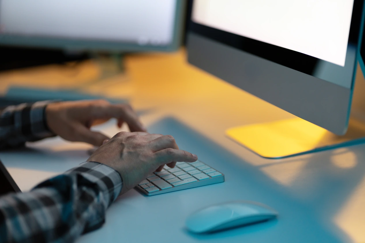 Dłonie programisty piszące na bezprzewodowej klawiaturze Apple w zaciemnionym biurze przy komputerze iMac.