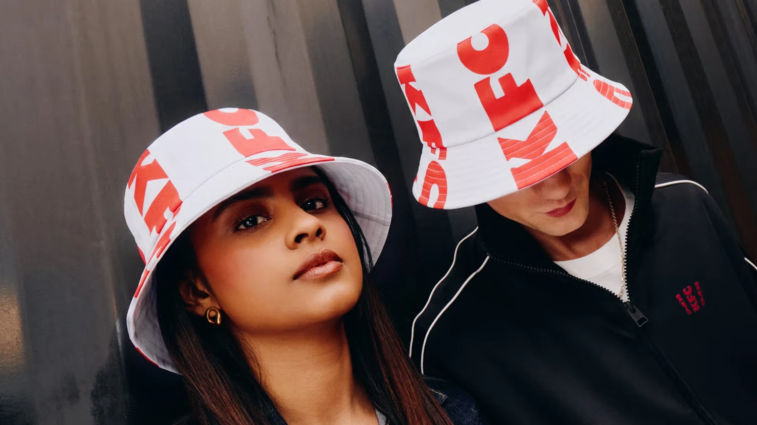 Dwoje modeli w białych kapeluszach typu bucket hat z czerwonym logo KFC, pozujących na tle ciemnej, prążkowanej metalowej ściany.