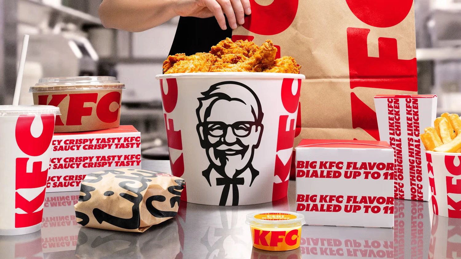Duży zestaw jedzenia KFC z kubełkiem chrupiących kawałków kurczaka, frytkami, burgerem w papierku, sosami i napojem w kubku, ułożony na metalowym blacie na tle torby papierowej z logo marki.