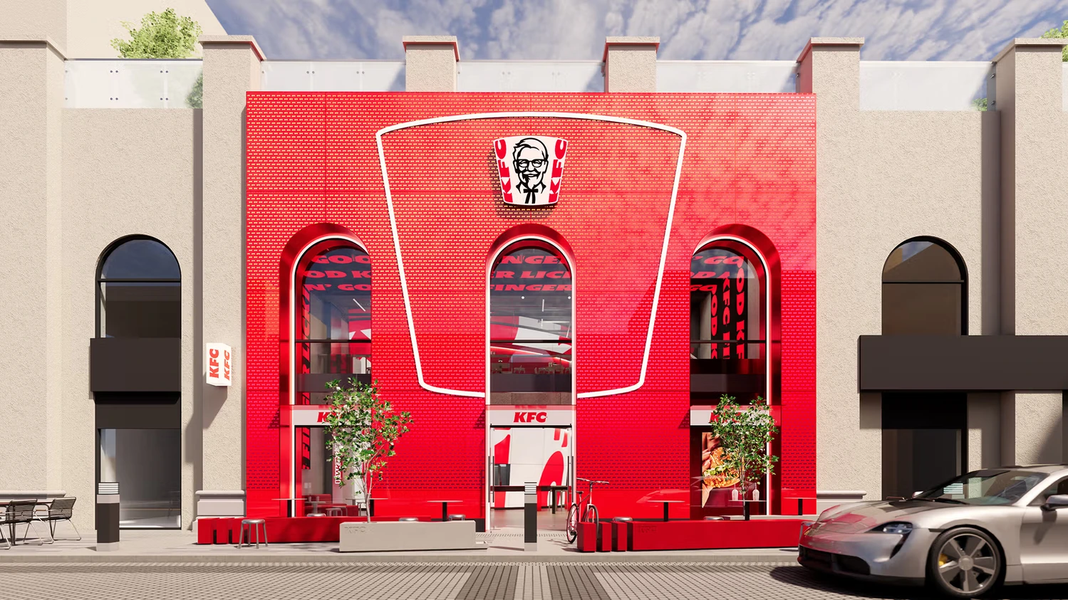 Nowoczesna fasada restauracji KFC wbudowana w jasną kamienicę. Czerwona, ceglana ściana frontowa ma kształt gigantycznego kubełka z logo Colonela Sandersa i dużymi szklanymi łukami okiennymi.