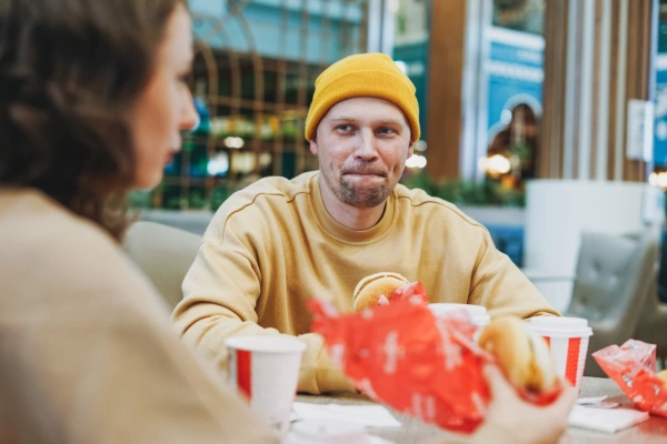 Mężczyzna w żółtej czapce i bluzie je burgera w restauracji szybkiej obsługi, rozmawiając z siedzącą naprzeciwko kobietą. Na stole widać papierowe kubki i czerwone opakowania po jedzeniu.