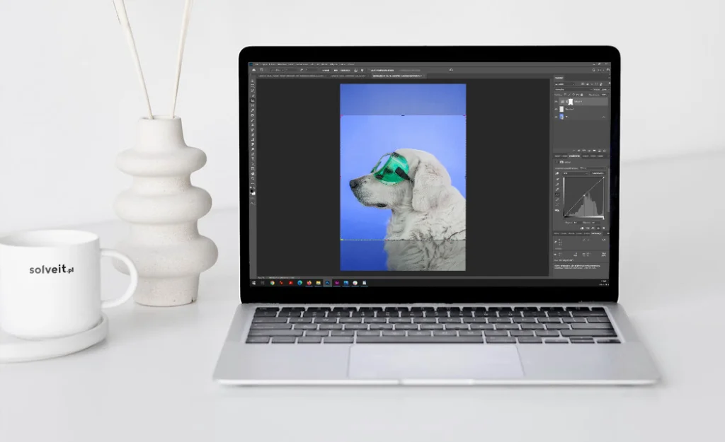 Otwarte srebrne laptop Apple MacBook Pro na białym biurku z wazonem, pałeczkami zapachowymi i kubkiem z logo solveit.pl. Ekran pokazuje interfejs Adobe Photoshop