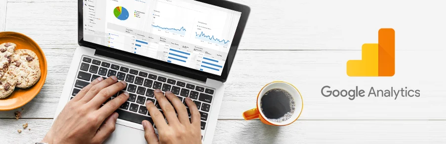 Widok z góry na laptop z otwartym panelem Google Analytics na ekranie. Obok kawa i ciasteczka na drewnianym stole