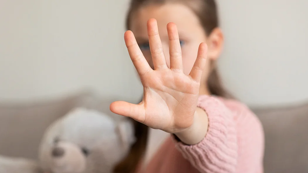 Otwarta dłoń małego dziecka wysunięta do przodu w geście „stop” lub „nie”. Twarz dziewczynki w różowym swetrze oraz leżący obok pluszowy miś są rozmyte i znajdują się w tle, poza głębią ostrości.