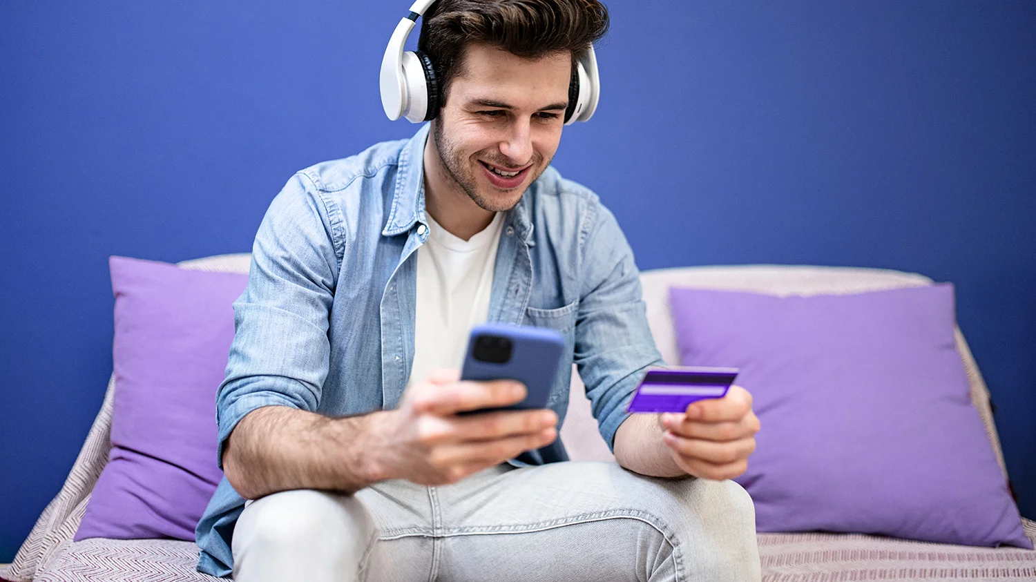 młody mężczyzna robi zakupy przez Internet Młody mężczyzna ubrany w jeans słucha muzyki i robi zakupy przez aplikację mobilną dzięki wprowadzaniu do niej danych z karty płatniczej