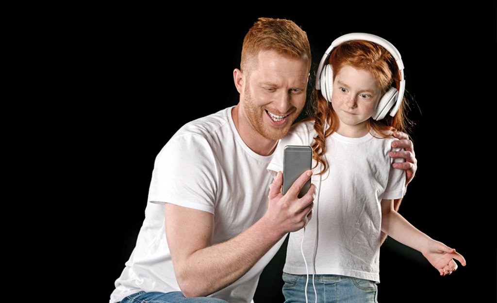 Uśmiechnięty, rudowłosy ojciec i mała córka w białych słuchawkach wspólnie oglądają wideo na smartfonie na czarnym tle. Kontrola rodzicielska w internecie.