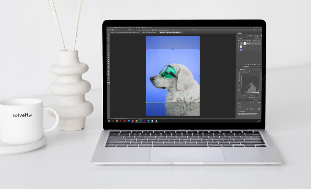 Otwarte srebrne laptop Apple MacBook Pro na białym biurku z wazonem, pałeczkami zapachowymi i kubkiem z logo solveit.pl. Ekran pokazuje interfejs Adobe Photoshop