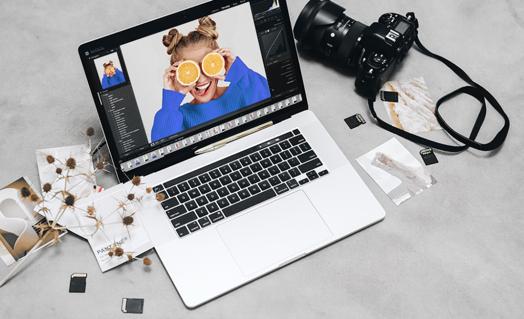 Ujęcie z góry przedstawiające laptop MacBook z otwartym zdjęciem edytowanym w programie Lightroom. Obok leży aparat fotograficzny Sigma, paski aparatowe, karty pamięci SD