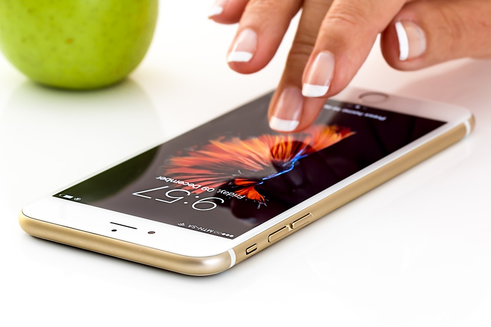 Bliskie ujęcie kobiecych dłoni z zadbanym francuskim manicure, dotykających ekranu złotego iPhone'a spoczywającego na białej powierzchni obok zielonego jabłka.
