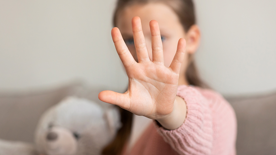 Otwarta dłoń małego dziecka wysunięta do przodu w geście „stop” lub „nie”. Twarz dziewczynki w różowym swetrze oraz leżący obok pluszowy miś są rozmyte i znajdują się w tle, poza głębią ostrości.