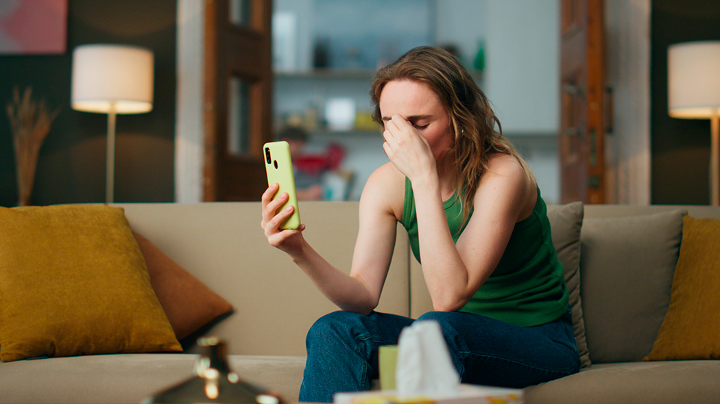 Zmartwiona kobieta w zielonym topie i jeansach siedzi na beżowej kanapie, trzymając w jednej ręce jasny smartfon, a drugą masując nasadę nosa i zasłaniając oczy w geście rezygnacji lub bólu głowy. Na stole przed nią widoczne jest pudełko z chusteczkami higienicznymi.