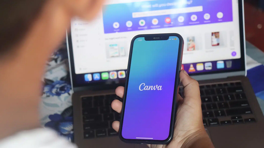 Mężczyzna trzyma w dłoni smartfon z uruchomionym fioletowo-niebieskim ekranem startowym aplikacji Canva. W tle, za telefonem, widoczny jest otwarty laptop z wyświetloną przeglądarkową wersją tego samego programu graficznego.