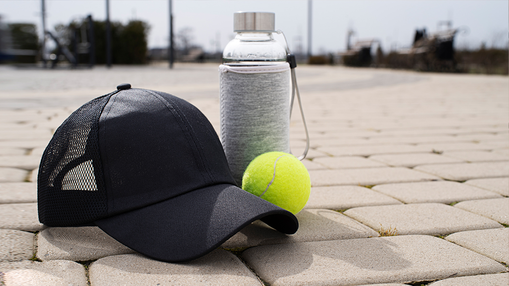 sportowe gadżety reklamowe czapka, piłka i butelka szklana na kostce brukowej niedaleko kortów tenisowych