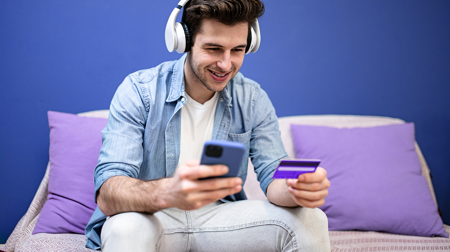 młody mężczyzna robi zakupy przez Internet Młody mężczyzna ubrany w jeans słucha muzyki i robi zakupy przez aplikację mobilną dzięki wprowadzaniu do niej danych z karty płatniczej