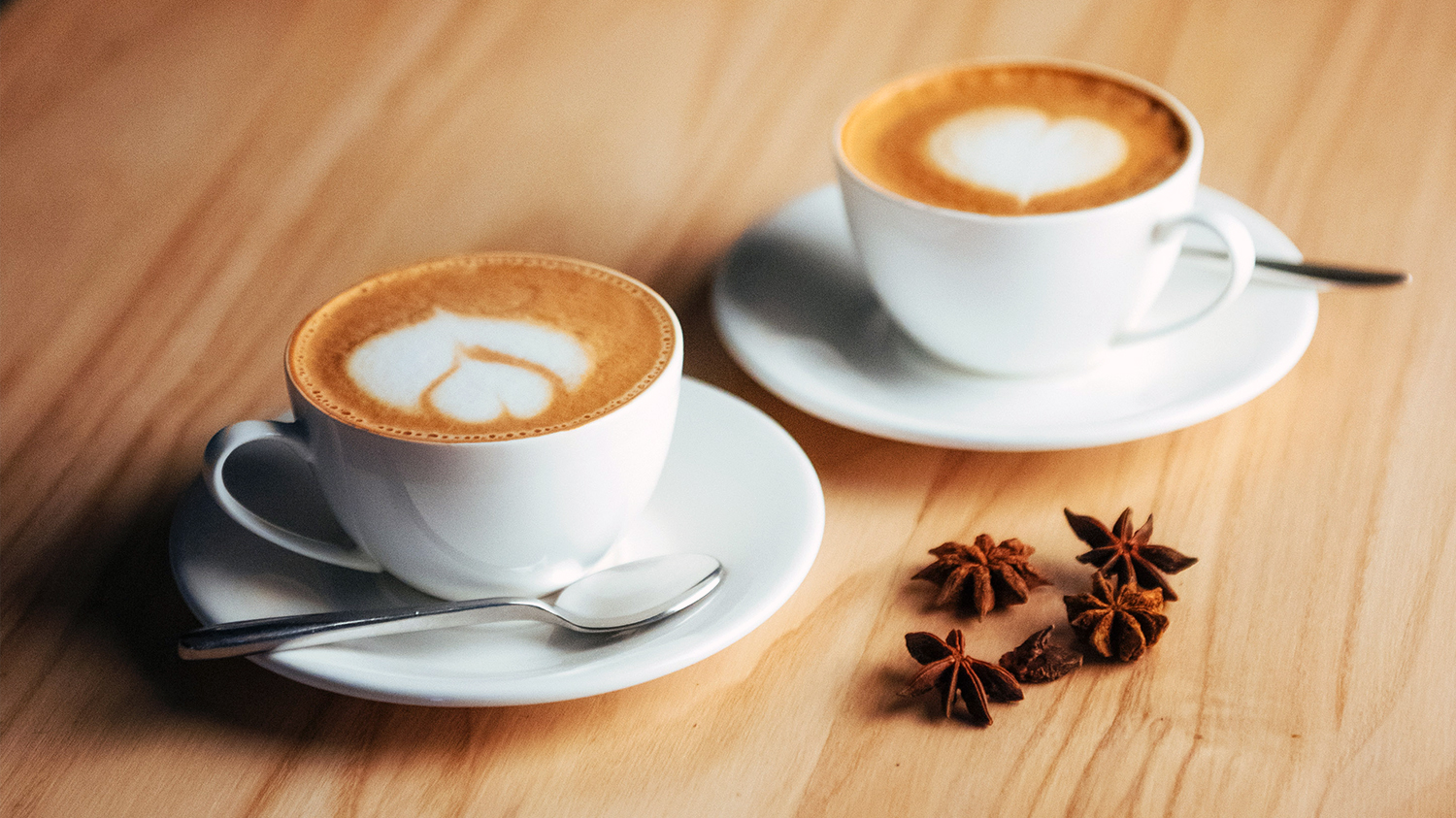 dwie filiżanki kawy Na jasnym drewnianym blacie stoją dwie filiżanki cappucino a obok nich leżą gwiazdki anyżu.